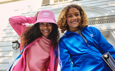 Two young women with curly hair, one wearing a pink hat and the other a blue jacket, are smiling and posing in front of a building with arched windows.