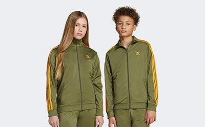 Two young people wearing olive green and yellow athletic jackets and pants stand against a plain white background.