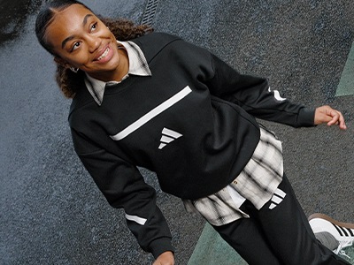 A young woman with curly hair wearing a black and white athletic jacket stands in front of a gray concrete wall, smiling confidently.