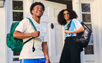 Two young people, a man and a woman, are standing in front of a building with windows. The man is wearing a white t-shirt and the woman is wearing a black top. They both have backpacks and appear to be smiling.