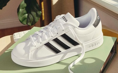 A pair of white Adidas sneakers with black stripes sits on a green platform, with a potted plant visible in the background.