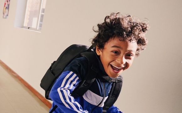 A young person with curly hair wearing a backpack and smiling brightly in what appears to be a hallway or indoor setting.