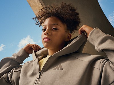 A young person with curly hair wearing a beige jacket stands against a bright blue sky.