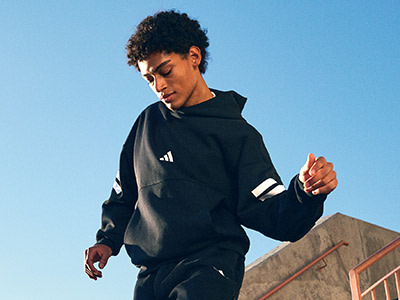 A young person wearing a black hooded sweatshirt stands against a clear blue sky, with a brick wall visible in the background.
