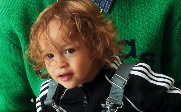 A young child with curly reddish-brown hair and a serious expression, wearing a black and white striped outfit, stands in front of a vibrant green background.