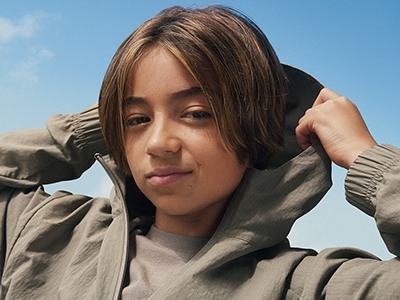 A young woman with long, dark hair wearing a beige hooded jacket stands against a clear blue sky.