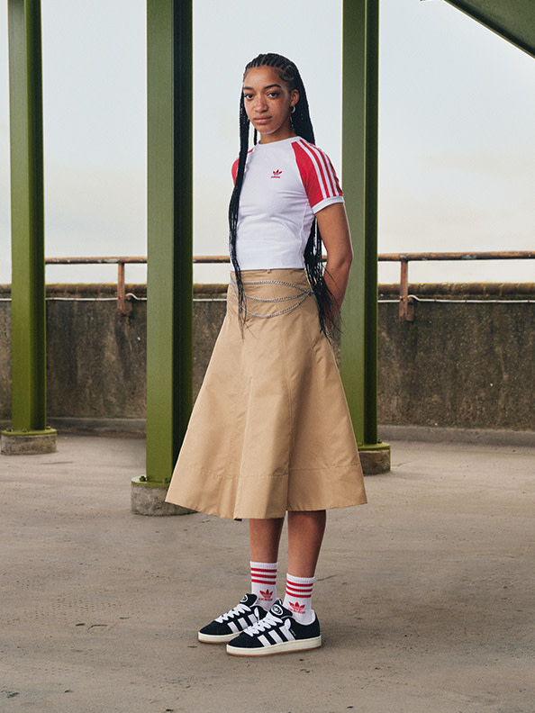 A young woman wearing a white top with red stripes, a beige skirt, and black and white sneakers stands in front of a concrete structure with a railing.