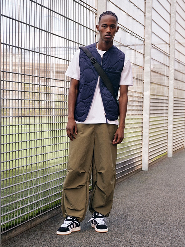 A young man wearing a navy blue vest, a white t-shirt, khaki cargo pants, and black and white sneakers stands in front of a metal mesh fence.