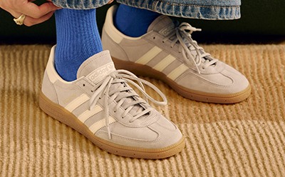 A pair of beige and blue sneakers with white stripes sit on a wooden floor, with a denim-clad leg visible in the foreground.