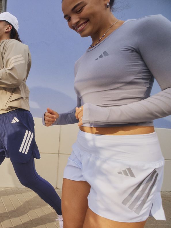 The image shows two individuals, one wearing a gray top and the other wearing navy blue shorts, standing in an outdoor setting with a blue sky in the background.
