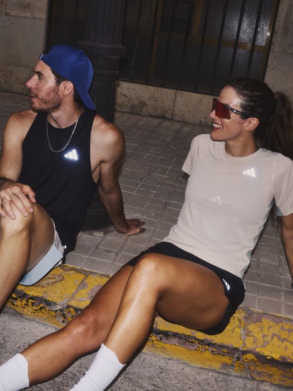 Two people, a man and a woman, are sitting on the ground in what appears to be an urban setting, with a brick wall visible in the background.