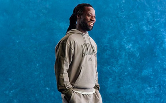 A man with dreadlocks wearing a beige hoodie stands against a vibrant blue background.