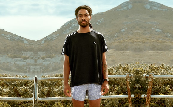 A man wearing a black t-shirt and shorts stands on a balcony overlooking a mountainous landscape in the background.