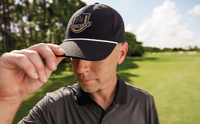 A person wearing a black baseball cap with a logo on it, standing in a grassy field with trees in the background.