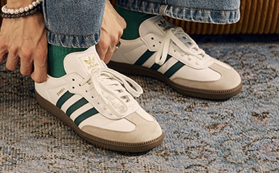 The image shows a pair of white and green Adidas sneakers on a patterned carpet.