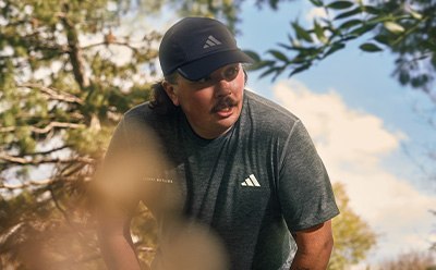 A man wearing an Adidas cap and shirt stands in a wooded area with trees and foliage in the background.