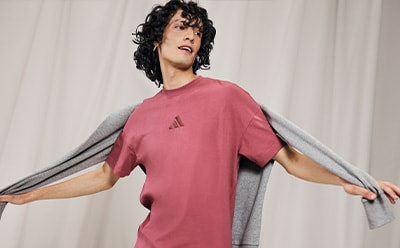 A person with curly dark hair wearing a pink t-shirt stands against a plain white background, their arms outstretched in a dramatic pose.