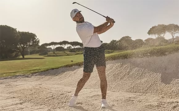 A person in athletic clothing is swinging a golf club on a grassy golf course surrounded by trees.