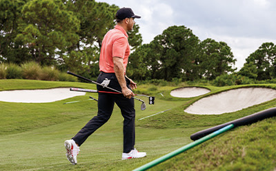A person in a pink shirt and black pants is standing on a golf course, surrounded by lush greenery and sand traps.