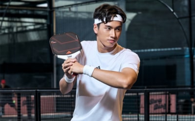 A young man in a white t-shirt is holding a tennis racket and standing in an outdoor sports facility with fencing and structures visible in the background.
