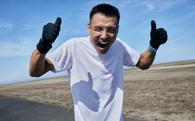 A man in a white t-shirt and black gloves is enthusiastically gesturing with his hands against a backdrop of a clear blue sky and a dry, sandy landscape.