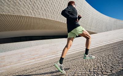A person in a dark jacket and green shorts stands on a paved surface in front of a curved, textured building against a clear blue sky.