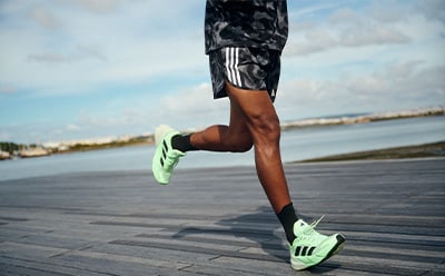 The image shows a person's lower body wearing patterned athletic shorts and bright green sneakers, running on a paved path with a body of water and buildings visible in the background.