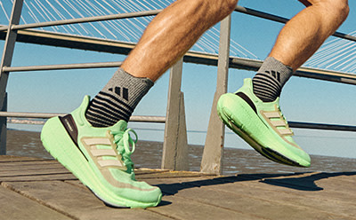 The image shows a pair of feet wearing bright green and white athletic shoes on a wooden platform against a backdrop of a blue sky and a suspension bridge.