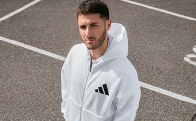 A man wearing a white hooded sweatshirt stands on a paved surface with painted lines, suggesting an athletic or sports-related setting.