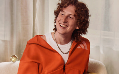 A smiling individual with curly red hair wearing a bright orange jacket sits in a room with white curtains in the background.