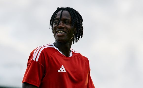 A person with dreadlocks wearing a red sports jersey stands against a cloudy sky background.