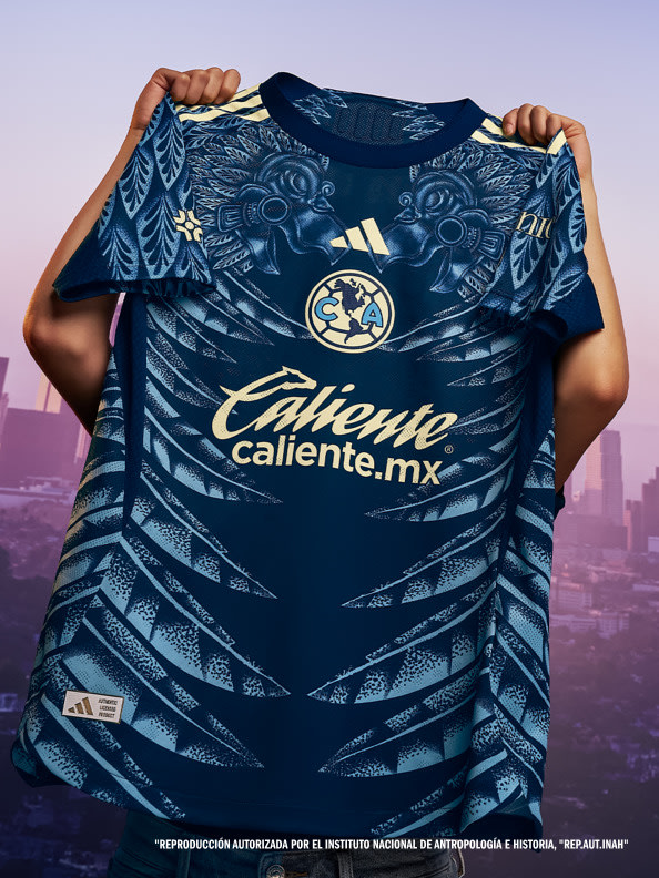 A person is holding up a navy blue and gold soccer jersey with a distinctive pattern and the "Caliente.mx" logo prominently displayed, set against a blurred urban skyline in the background.
