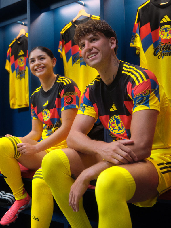 Two people wearing colorful soccer jerseys with the Club América logo sit together in what appears to be a locker room or team facility.