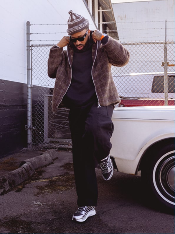 A person wearing a brown fur coat, black pants, and sneakers stands in front of a chain-link fence and a parked vehicle.