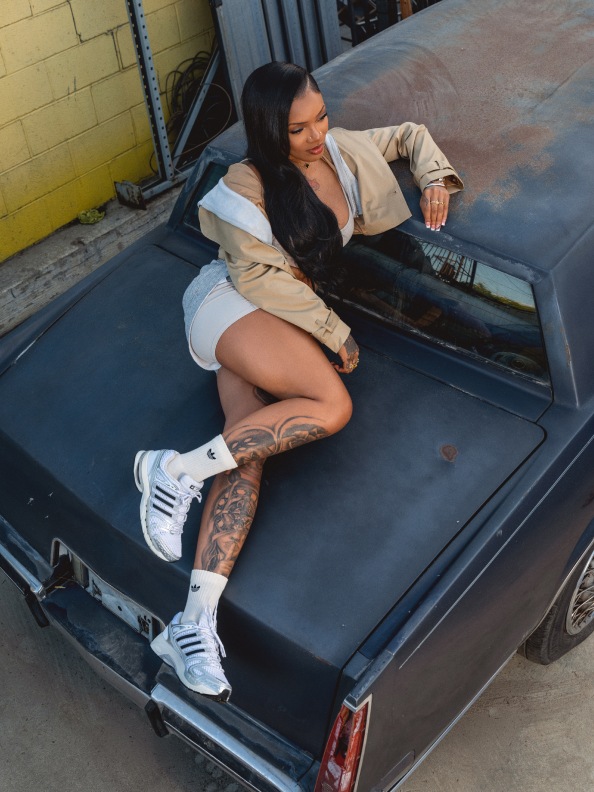 A woman with long dark hair is sitting on the back of a large black vehicle, wearing a beige jacket and white sneakers, with visible tattoos on her legs.