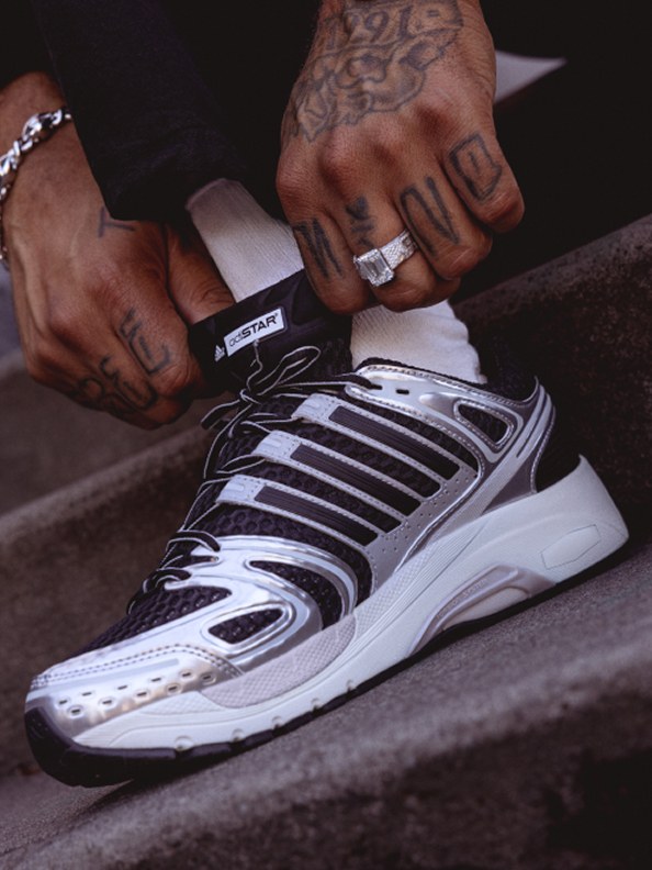 The image shows a pair of hands with tattoos adjusting the laces of a white and black athletic shoe on a dark surface.