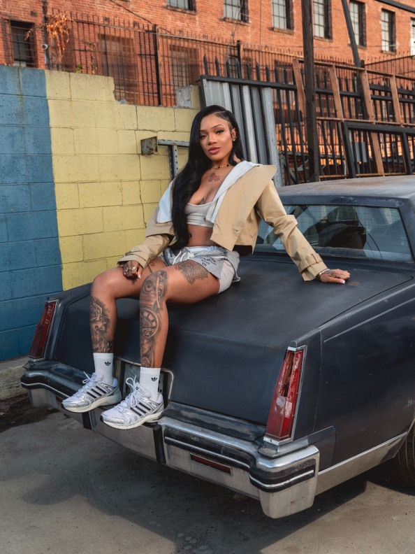 A young woman with long dark hair is sitting on the back of a large black vehicle, wearing a light-colored jacket and shorts, and displaying tattoos on her legs.