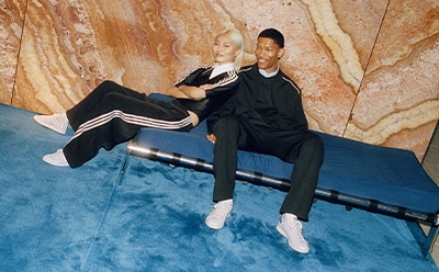 Two individuals, a man and a woman, are seated on a bench against a textured, orange-toned wall background.