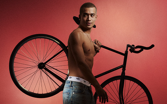 A shirtless young man stands next to a vintage bicycle against a vibrant red background.