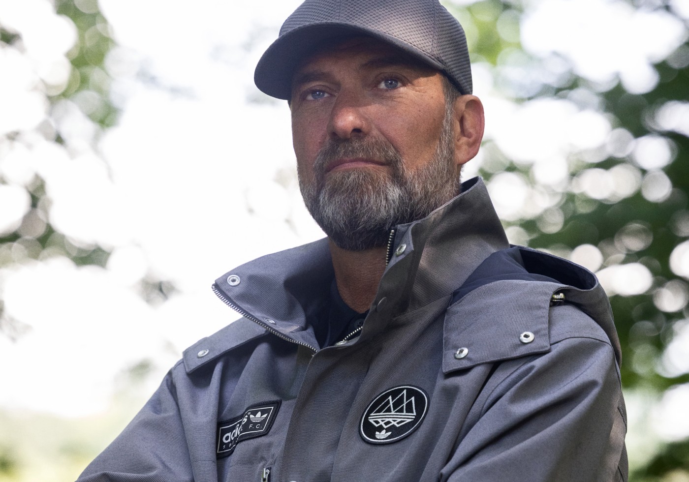 Jürgen Klopp wears a gray jacket and cap from the adidas SPEZIAL collection, standing in a sunlit forest.