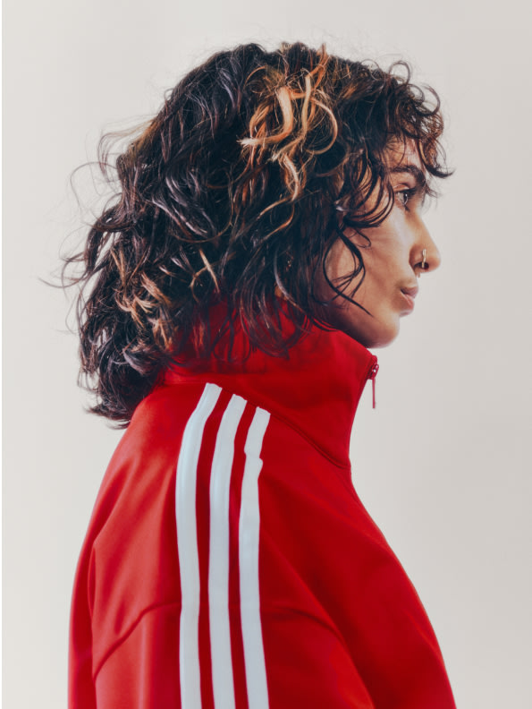 A portrait of a woman with brown hair wearing a red adicolor tracksuit in front of a white backdrop. 