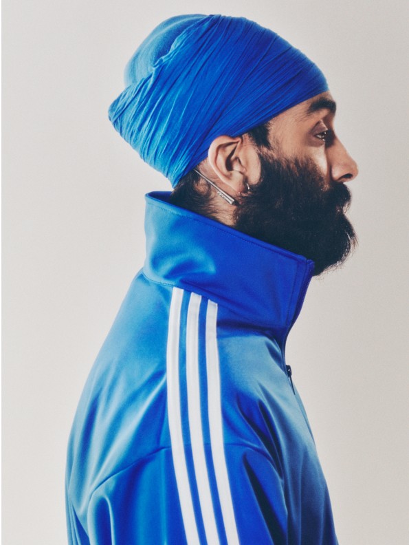 A portrait of a man in a blue turban and a blue adicolor tracksuit standing in front of a white backdrop. 