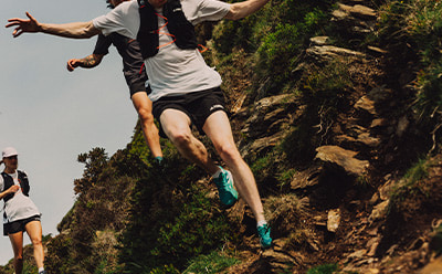 A person is jumping off a rocky cliff, surrounded by lush greenery and a dramatic landscape in the background.