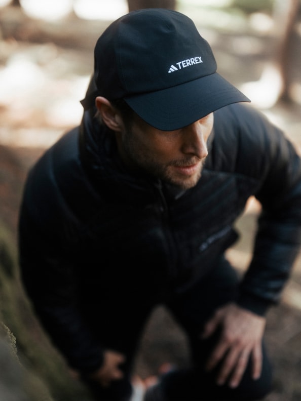 Eine Person mit schwarzer adidas Kappe und Jacke hockt sich in einem bewaldeten Outdoor-Setting zusammen.