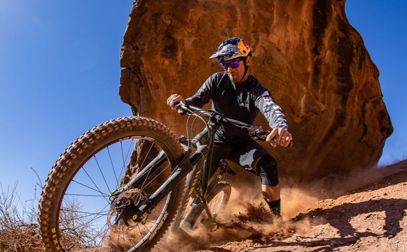 En mountainbiker kører ned ad en stenet, støvet sti mod en klar blå baggrund.