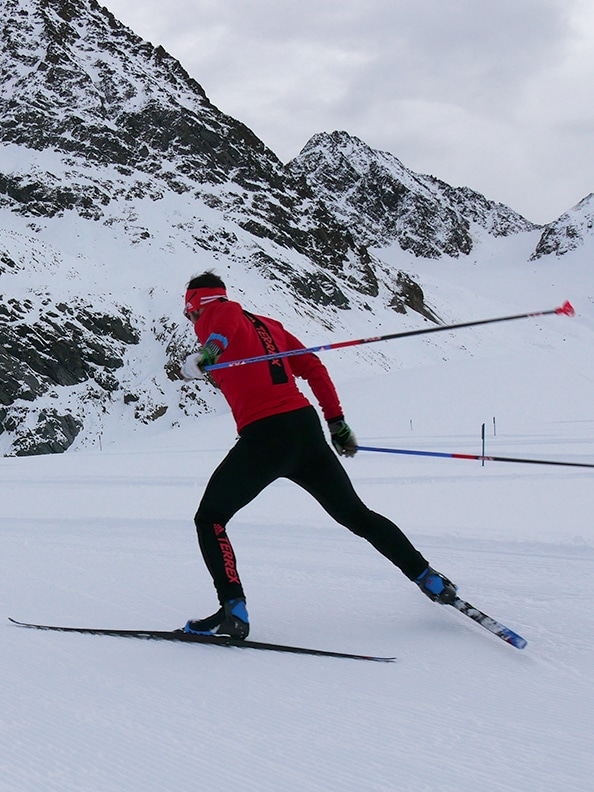 Una esquiadora con chaqueta roja esquia por una pendiente cubierta de nieve sobre un fondo de montañas escarpadas y cubiertas de nieve.