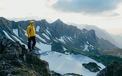 Una persona con una chaqueta amarilla se coloca de pie en un acantilado rocoso con vistas a un paisaje montañoso con picos cubiertos de nieve y un lago debajo.