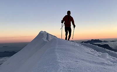 A person in a red jacket stands on a snowy ridge, with mountains silhouetted against a colorful sunset sky in the background.