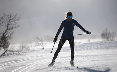 A person in a black ski suit is skiing on a snowy trail, with bare trees and a cloudy sky in the background.