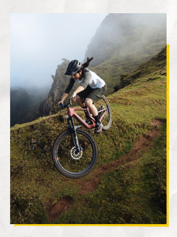Uma pessoa a pedalar numa BTT por uma encosta íngreme e herbácea numa paisagem enevoada e montanhosa.